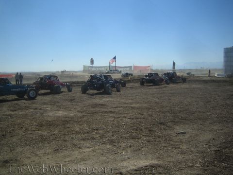 1990 Ford Bronco - Several off-road buggies and trucks staged on a dusty dirt course with an American flag and banners in the hazy distance.