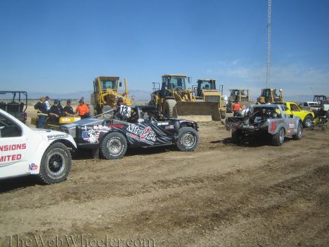 1990 Ford Bronco - lineup of off-road race trucks including number 13 sits on a dirt staging area with bulldozers and a clear blue sky behind.