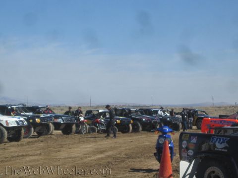 1990 Ford Bronco - long row of off-road race trucks and small trophy karts staged on a dirt lot under a hazy sky, with an orange cone in the foreground.