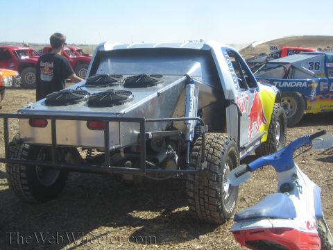 1990 Ford Bronco - silver off-road truck fitted with four rear-mounted cooling fans sits beside a Red Bull-liveried race truck on a dirt course.