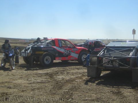 1990 Ford Bronco - red and black number 86 off-road race truck staged on a dirt course beside a parked dirt bike and other race vehicles.