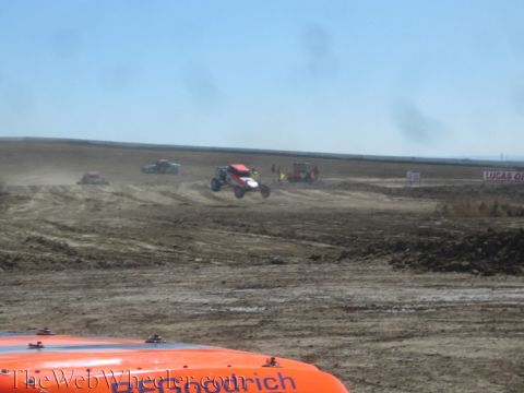 1990 Ford Bronco - off-road buggy catches air over a jump on a dusty dirt race track as other vehicles and banners line the course.