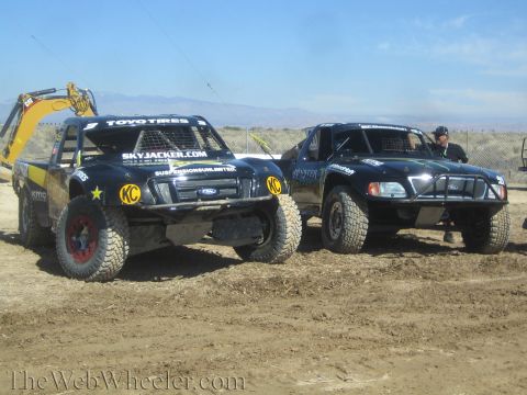 1990 Ford Bronco - Two black Ford off-road race trucks with Toyo Tires and Skyjacker graphics parked side by side on a dirt lot with mountains in the distance.