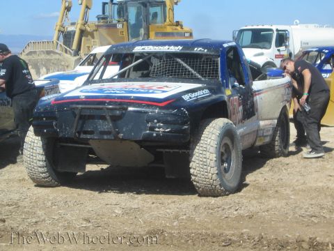 1990 Ford Bronco - dark blue BFGoodrich-sponsored off-road race truck sits on a dirt lot as a crew member leans into the cab with bulldozers behind.