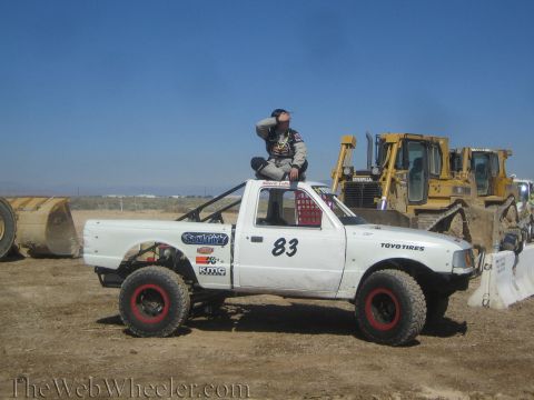1990 Ford Bronco - white number 83 Ford Ranger off-road race truck on a dirt lot with a man seated on the roof and Caterpillar bulldozers behind.