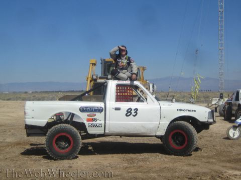 1990 Ford Bronco - white off-road race pickup truck marked number 83 sits on a dirt course as a helmeted driver stands looking out over the desert.
