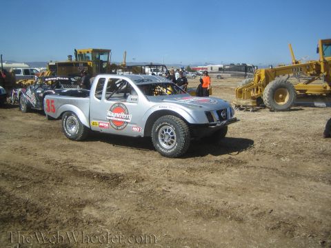 1990 Ford Bronco - silver off-road race truck numbered 35 covered in sponsor decals is parked on a dirt lot beside heavy construction equipment.