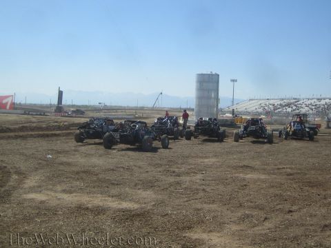 1990 Ford Bronco - row of open-wheel off-road race buggies lines up on a dusty dirt track with grandstands and a water tank behind them.