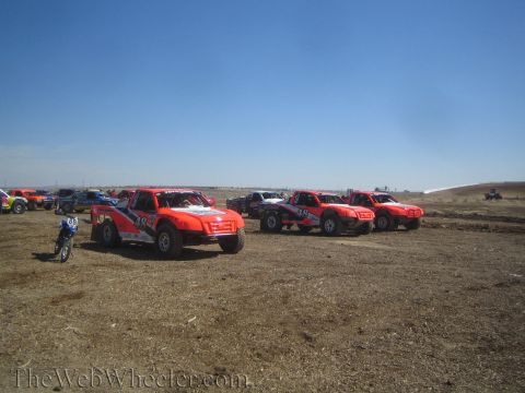 1990 Ford Bronco - Several orange and red off-road race trucks are staged on a bare dirt lot under a clear blue sky, with a small motorcycle nearby.