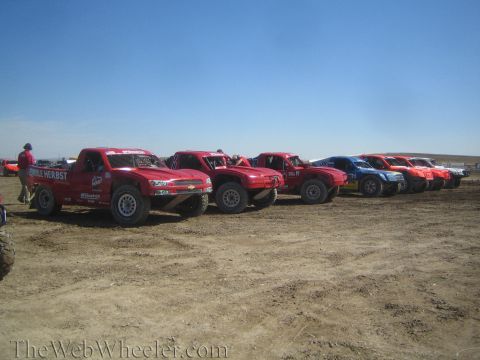 1990 Ford Bronco - long row of red and multicolored off-road race trucks is lined up on a dirt staging area as a crew member stands at left.