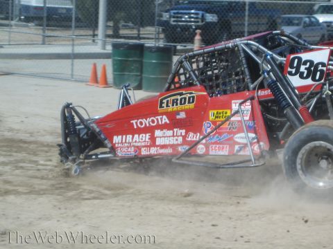 1990 Ford Bronco - red Toyota-badged off-road tube-chassis buggy slides through loose dirt, kicking up dust alongside number 936 near a track fence.