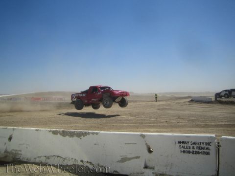 1990 Ford Bronco - dark red off-road race truck flies through the air off a jump on a hazy, dusty desert track behind a concrete barrier.
