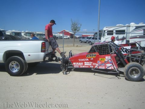 1990 Ford Bronco - man stands in the bed of a white dually pickup loading a red Toyota-badged off-road buggy in a dusty paddock area.
