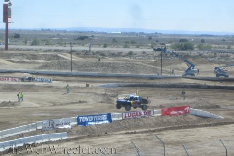 1990 Ford Bronco - off-road race truck catches air over a jump on a wide dirt track lined with sponsor banners and tall blue cranes in the distance.