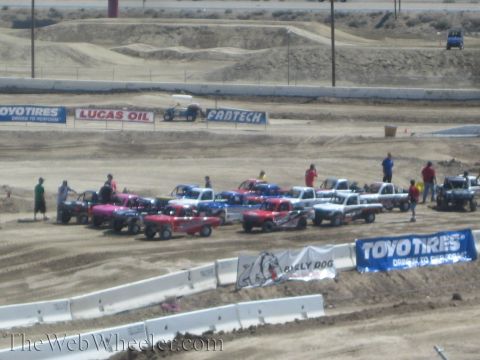 1990 Ford Bronco - cluster of colorful off-road race trucks is staged on a dirt track lined with Toyo Tires and Lucas Oil banners, seen from the stands.