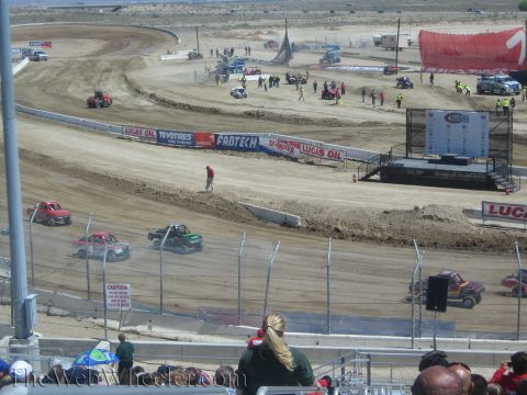 1990 Ford Bronco - Off-road race trucks circulate a banked dirt oval track ringed with Lucas Oil and Fabtech banners, viewed over spectators in the stands.