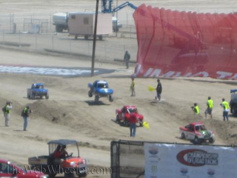 1990 Ford Bronco - Blue and pink off-road race trucks run a sandy track as flaggers in safety vests work near a large red Championship Off-Road banner.