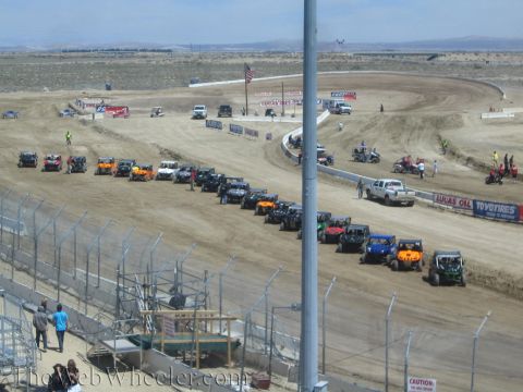 1990 Ford Bronco - Rows of off-road buggies and trucks are staged across a wide dirt track beneath a US flag, viewed from the grandstands.