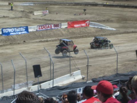 1990 Ford Bronco - Two side-by-side UTVs race close together on a dirt track lined with Toyo Tires and Lucas Oil banners as spectators watch in the foreground.
