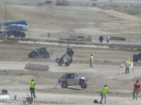 1990 Ford Bronco - off-road UTV is flipped onto its side on a dusty track while a yellow-vested safety crew and other racing vehicles stand nearby.