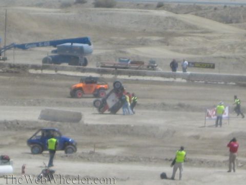 1990 Ford Bronco - Crew members surround a rolled UTV on a dusty track as an orange off-road buggy and a blue buggy pass nearby with a crane behind.