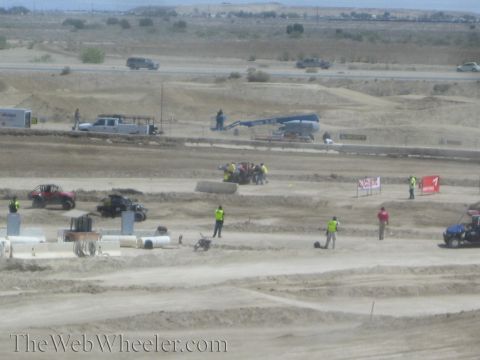 1990 Ford Bronco - Yellow-vested safety crew clear debris on a wide, dusty off-road track with a few buggies and a blue crane in the background.