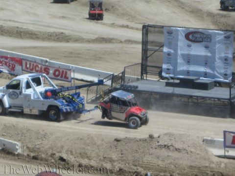 1990 Ford Bronco - white Lucas Oil tow truck pulls a red off-road UTV off the dirt track beside a sponsor stage backdrop.