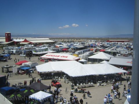 1990 Ford Bronco - elevated view shows a crowded off-road race paddock packed with white tents, sponsor trailers and Lucas Oil canopies under a blue sky.