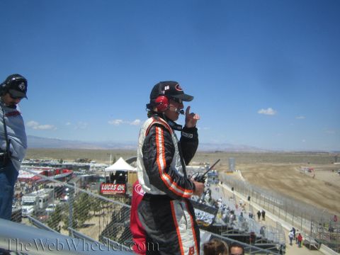 1990 Ford Bronco - man in a black-and-orange racing suit holds a radio and signals while standing on an elevated platform overlooking a desert race venue.
