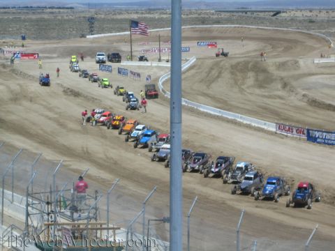 1990 Ford Bronco - long line of off-road buggies and trucks is staged along a dirt oval track beneath a US flag, viewed from the grandstands.