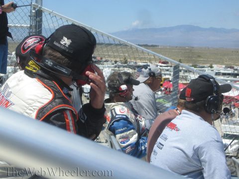 1990 Ford Bronco - Drivers and crew in helmets and radio headsets gather behind a chain-link track fence overlooking the paddock and distant mountains.