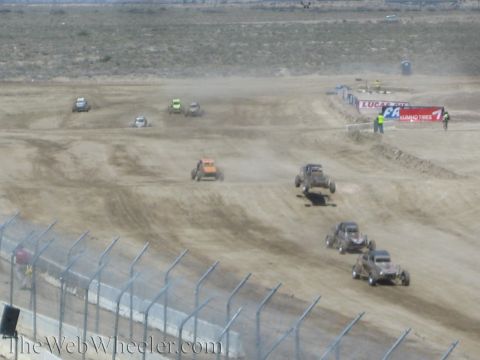 1990 Ford Bronco - pack of off-road short-course race trucks, one airborne, kicks up dust on a dirt circuit lined with sponsor banners.