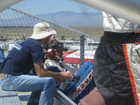 1990 Ford Bronco - Spectators and crew in caps and racing suits sit in grandstands overlooking a desert racetrack.