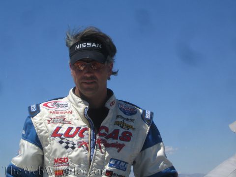 1990 Ford Bronco - man wearing a white and blue Lucas-branded racing suit and a Nissan visor stands outdoors under a blue sky.