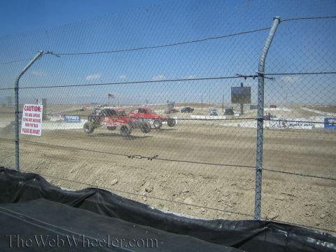 1990 Ford Bronco - Red off-road race trucks run side by side on a dusty dirt track viewed through a chain-link safety fence.