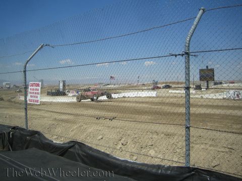 1990 Ford Bronco - red off-road race truck rounds a dirt track behind a chain-link fence bearing a red caution sign.