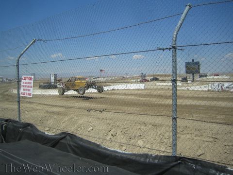 1990 Ford Bronco - yellow tube-frame off-road race truck runs a dirt course viewed through a chain-link safety fence.
