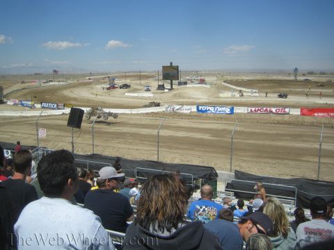 1990 Ford Bronco - crowd in grandstands watches off-road race trucks circle a wide desert dirt track with sponsor banners.