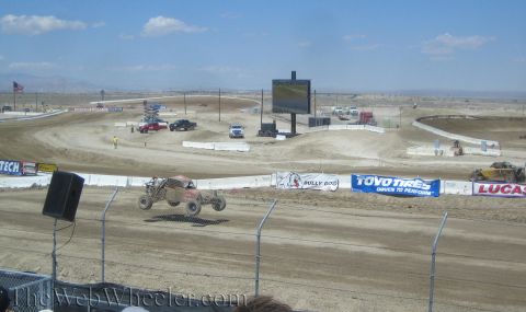 1990 Ford Bronco - tube-frame off-road race truck catches air on a desert dirt track near a large video screen.