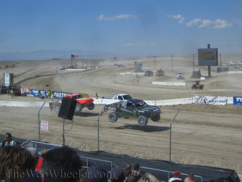 1990 Ford Bronco - black Monster-branded truck and a red truck race over jumps on a dusty desert short-course track.