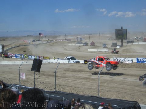 1990 Ford Bronco - orange and white number-86 off-road race truck launches off a jump on a desert dirt course before spectators.