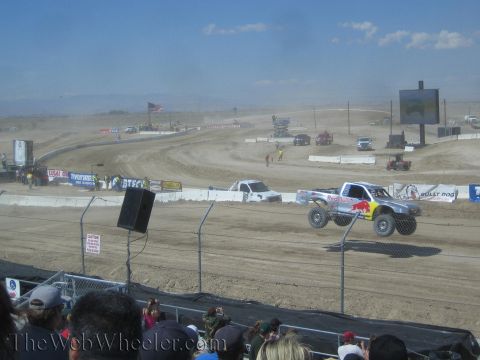 1990 Ford Bronco - silver Red Bull off-road race truck and a white truck speed along a dusty desert dirt course before a crowd.