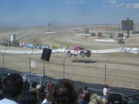 1990 Ford Bronco - red and white off-road race truck catches air on a desert dirt course as a crowd looks on.