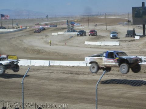 1990 Ford Bronco - Two off-road race trucks power through a dusty dirt turn viewed past a chain-link safety fence.
