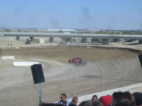 1990 Ford Bronco - red off-road race truck kicks up dust on a dirt track with a freeway overpass visible behind it.