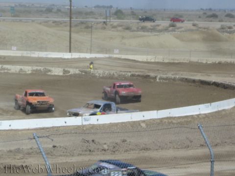 1990 Ford Bronco - Orange, gray and red off-road race trucks fight through a desert dirt turn near a highway.