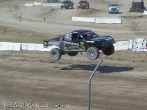 1990 Ford Bronco - black Monster-branded off-road race truck catches air over a jump on a desert dirt course.