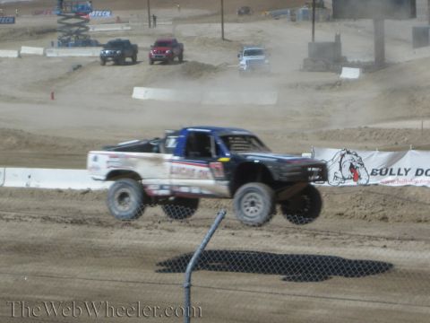 1990 Ford Bronco - white and black Lucas Oil off-road race truck flies over a jump beside a Bully Dog sponsor banner.