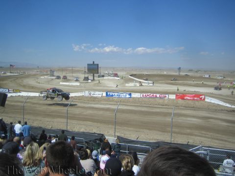 1990 Ford Bronco - off-road race truck runs a wide desert dirt course lined with sponsor banners as a grandstand crowd watches.