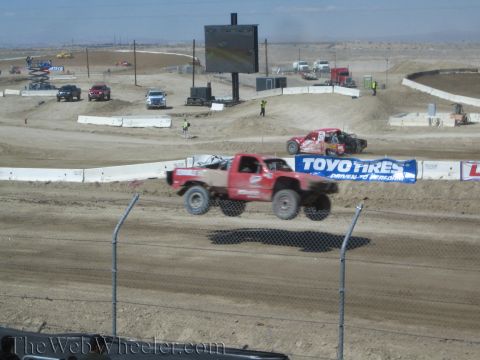 1990 Ford Bronco - red number-2 off-road race truck launches off a jump on a desert dirt track beside a Toyo Tires banner.